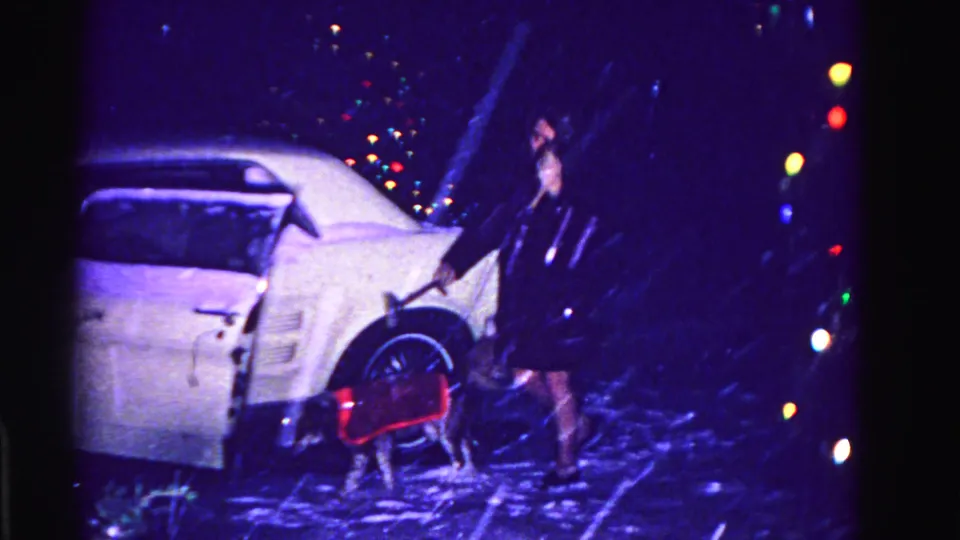 Woman in dark coat scraping snow from a white car door at night, small dog in red coat beside her, circa 1962 Hagerstown Maryland, multicolored Christmas lights glowing, 8mm film.