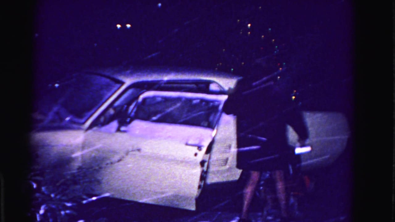 Woman in dark dress opens door of white American sedan at night in falling snow.