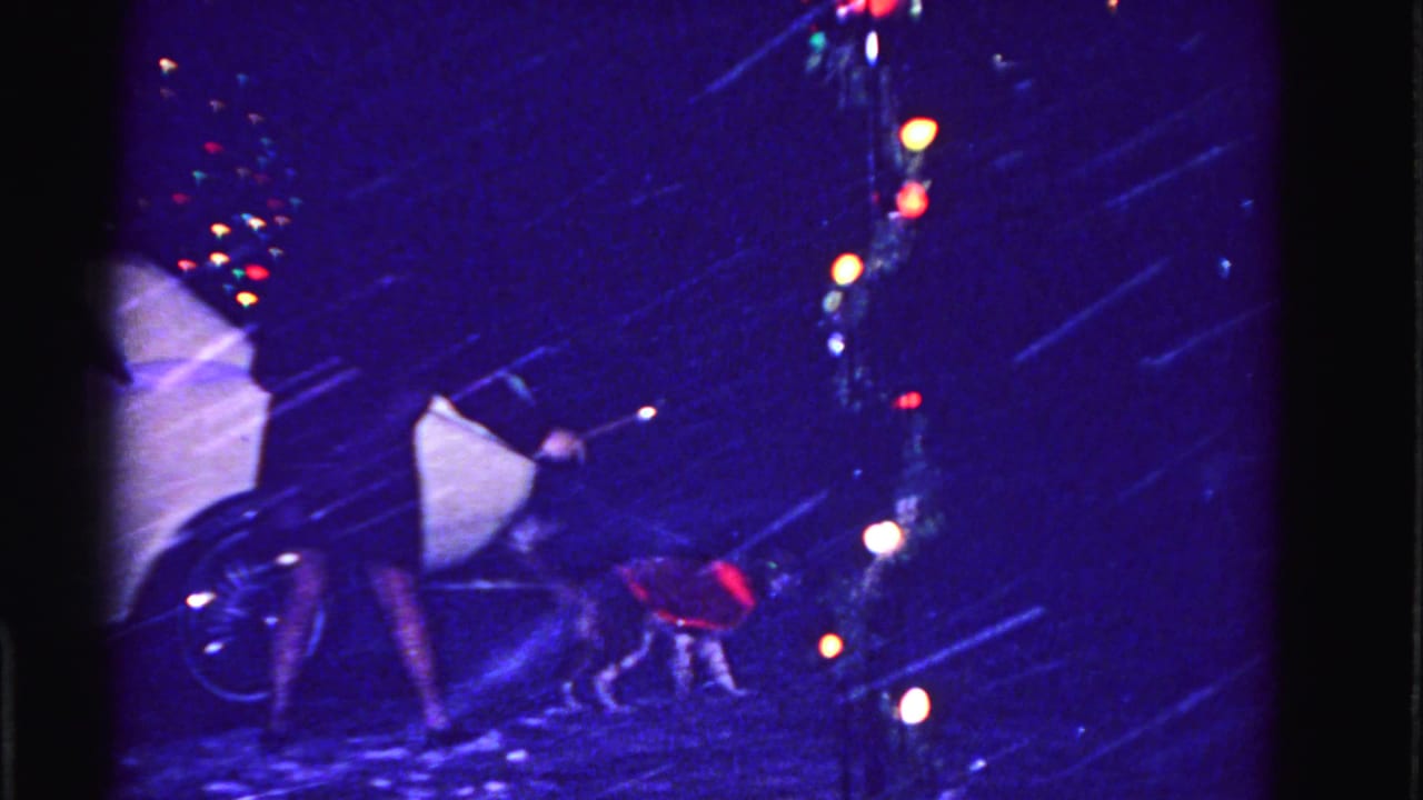 Woman bends toward small dog in red vest beside a classic car as colorful Christmas lights glow in the background during snowfall.