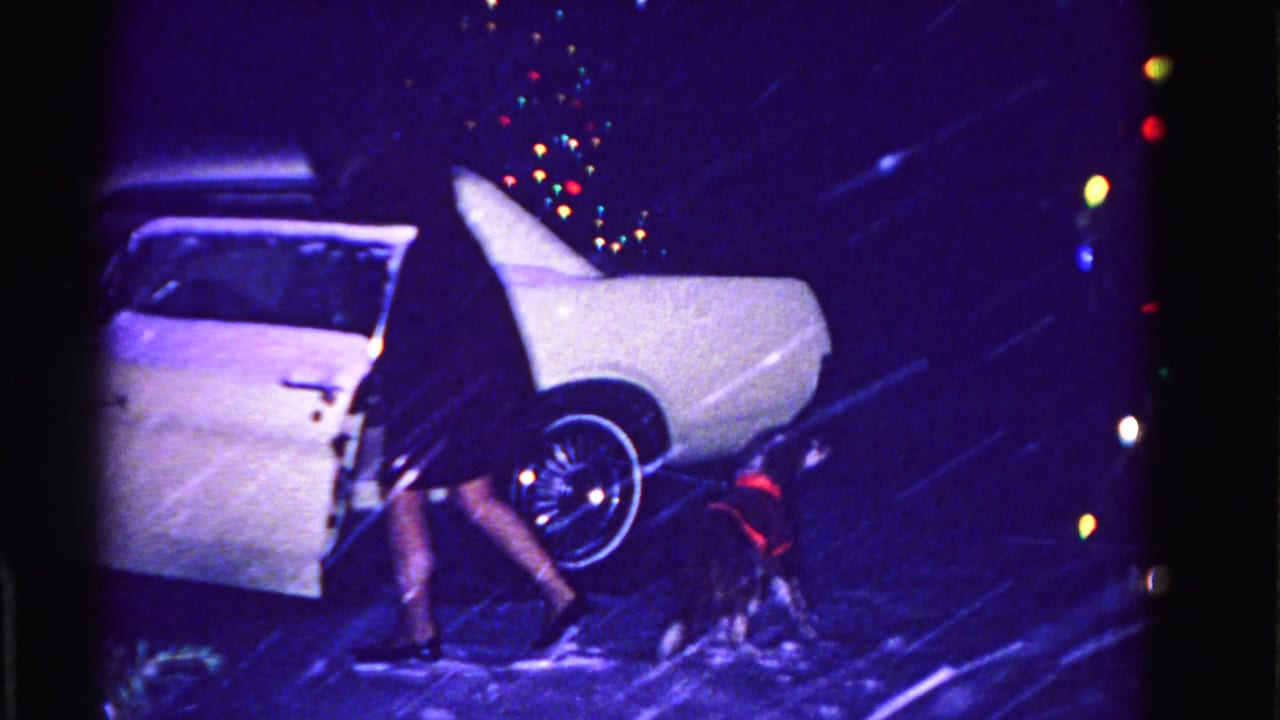 Woman in dark dress beside a white classic American car at night, small red-vested dog near rear wheel, multicolored holiday lights out of focus in background.