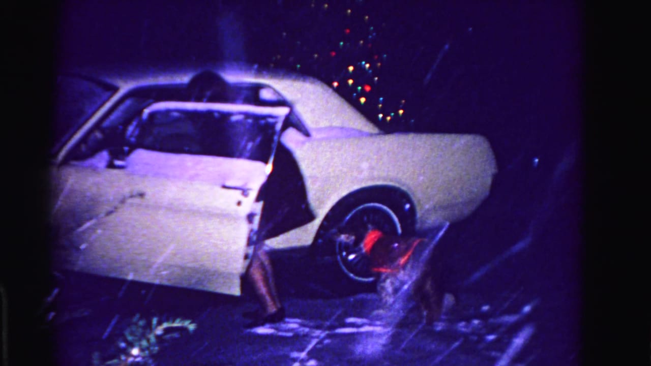 Woman preparing to enter white early-1960s American sedan at night, wet pavement and colorful holiday-light bokeh; Hagerstown, Maryland, circa 1962, 8mm film.