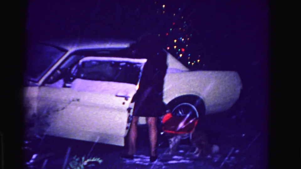 Woman at open driver's door of white early-1960s American sedan, multicolored holiday-light bokeh overhead, wet concrete; Hagerstown, Maryland, circa 1962, 8mm.