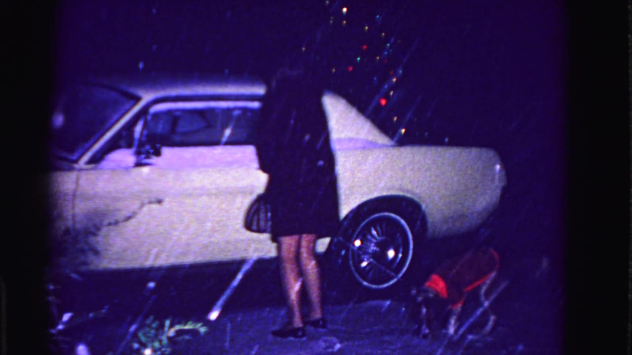 Lower body of a woman in dark skirt and heels alongside a white early-1960s car on a snowy night; small dog in red coat and bokeh Christmas lights visible, Hagerstown MD ca. 1962.