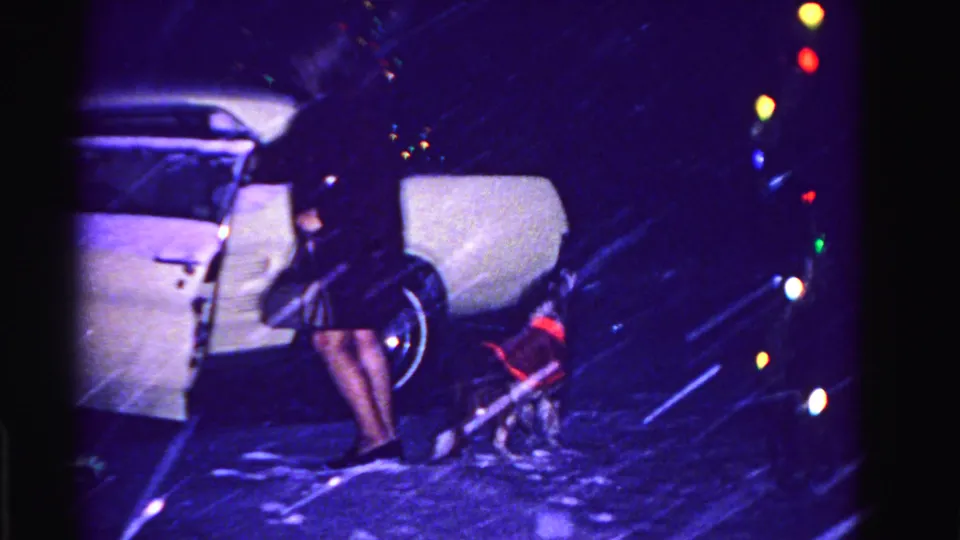 Woman in dark skirt boarding a white early-1960s car in heavy snow; string of round multicolored Christmas lights clearly visible at right, small dog near the car, Hagerstown MD ca. 1962.