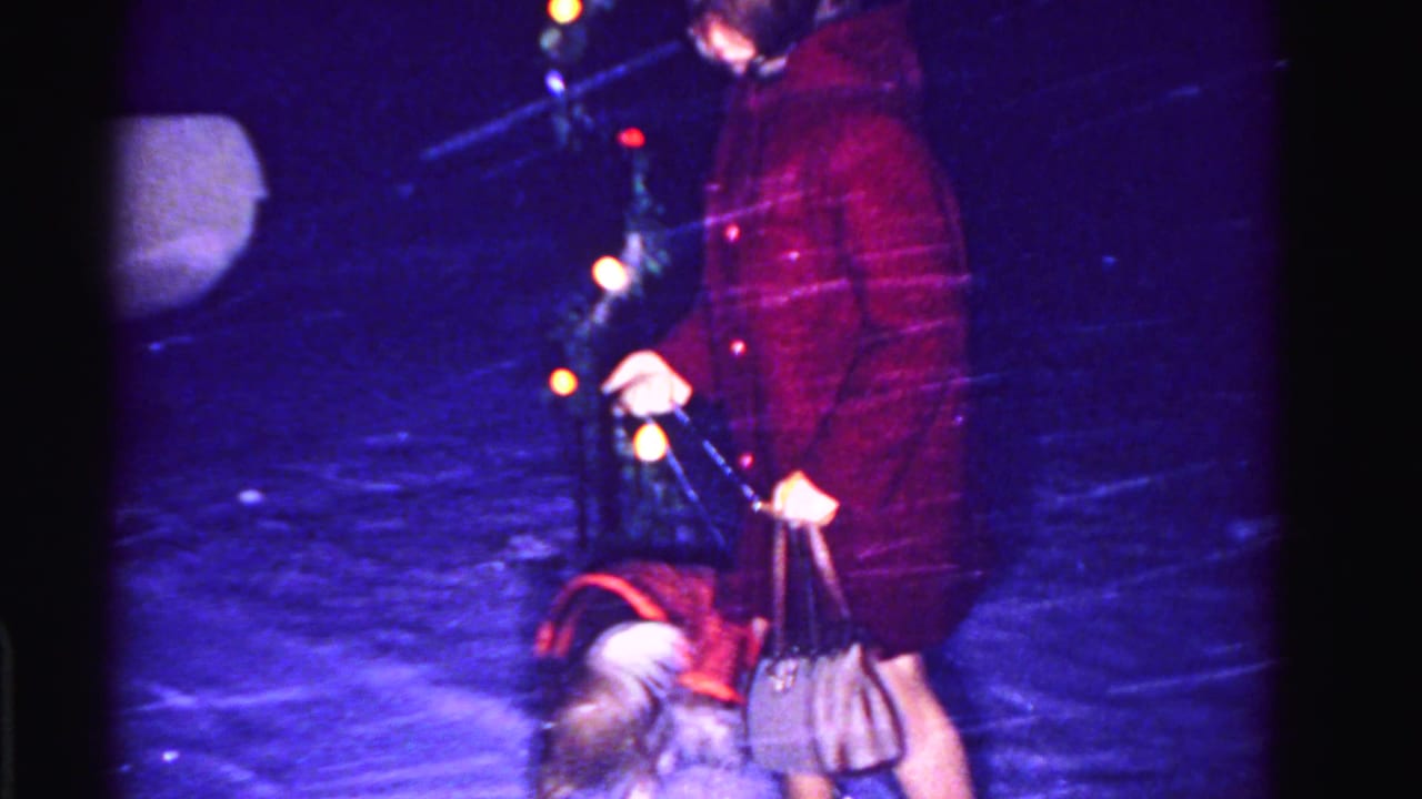 Close mid-shot of woman in dark red coat and handbag with small dog in falling snow, multicolor Christmas lights at right, circa 1962 8mm home movie.
