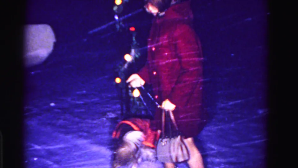 Close mid-shot of woman in dark red coat and handbag with small dog in falling snow, multicolor Christmas lights at right, circa 1962 8mm home movie.