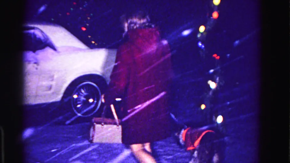 Woman in dark red coat walks a small dog past a large white American car in a snowy suburban night, multicolor Christmas lights at right, circa 1962.