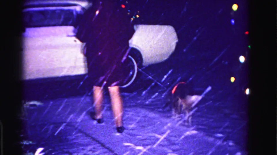 Woman in dark coat and heels walks away from camera with a small dog on leash through falling snow, white American car and multicolor Christmas lights behind, circa 1962.