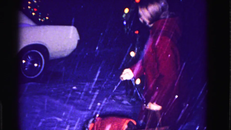 Woman in red coat outdoors in snowstorm at night, 1960s-era light-colored automobile and colored string lights visible, circa 1962 8mm footage.