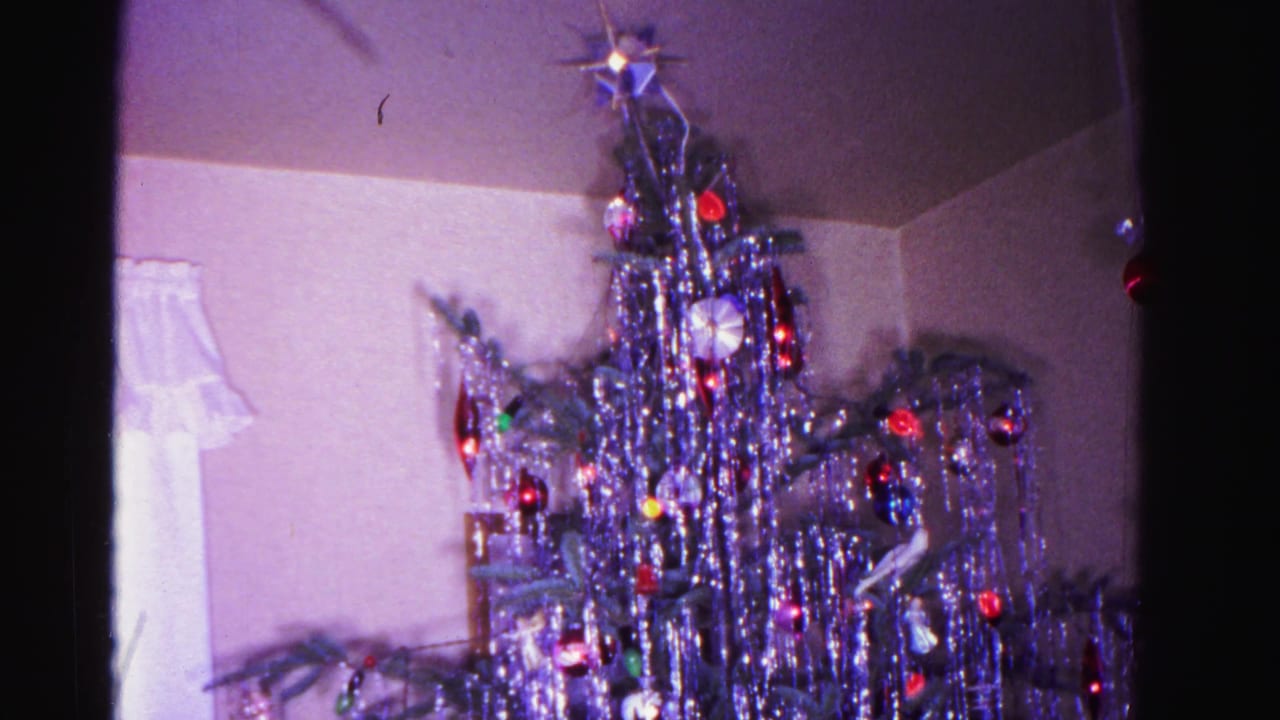 1962 Hagerstown, Maryland living room: silver aluminum tinsel Christmas tree with star topper, colored ornaments, and wrapped gifts at the base; partial figure at left edge; 8mm color home movie.