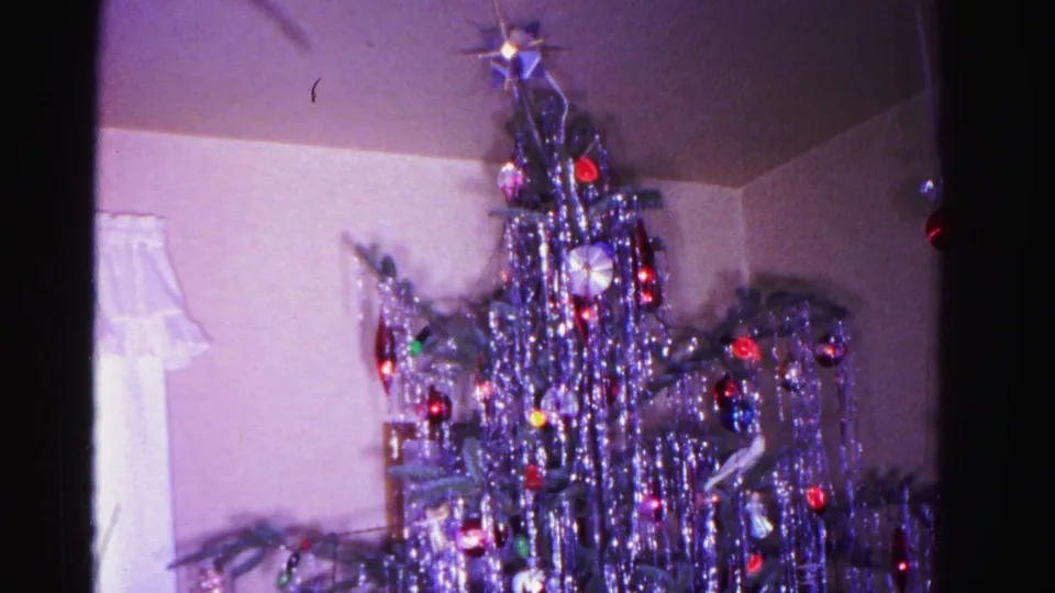 1962 Hagerstown, Maryland living room: silver aluminum tinsel Christmas tree with star topper, colored ornaments, and wrapped gifts at the base; partial figure at left edge; 8mm color home movie.