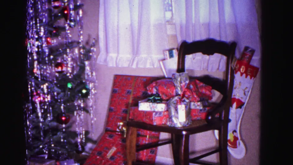 8mm home movie frame from 1962 showing wrapped gifts and a Christmas stocking on a wooden chair, with a tinseled tree beside a sheer-curtained window, Hagerstown, Maryland.