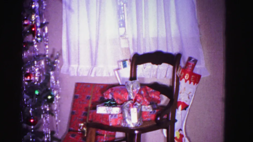 8mm home movie still from 1962 showing wrapped gifts and a Christmas stocking on a chair beside a tinseled tree in a domestic living room, Hagerstown, Maryland.