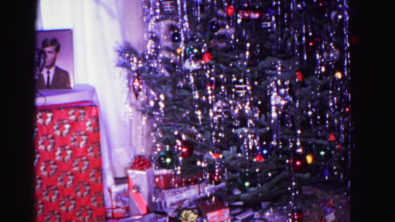 8mm home movie frame circa 1962: decorated Christmas tree with icicle tinsel, multicolored ornament lights, framed portrait of a man at left, and wrapped gifts beneath.