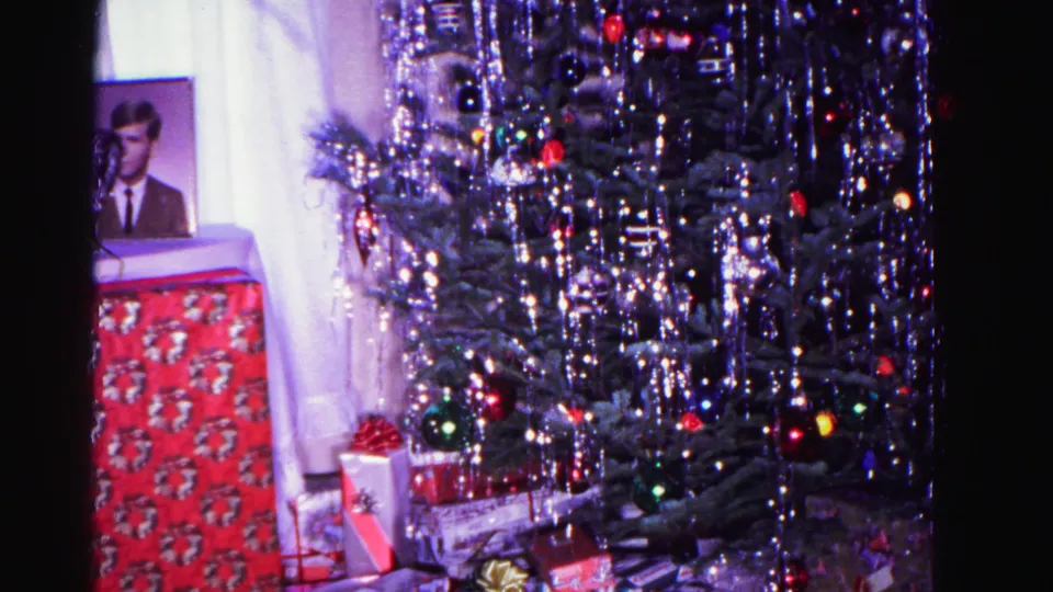 8mm home movie frame circa 1962: decorated Christmas tree with icicle tinsel, multicolored ornament lights, framed portrait of a man at left, and wrapped gifts beneath.