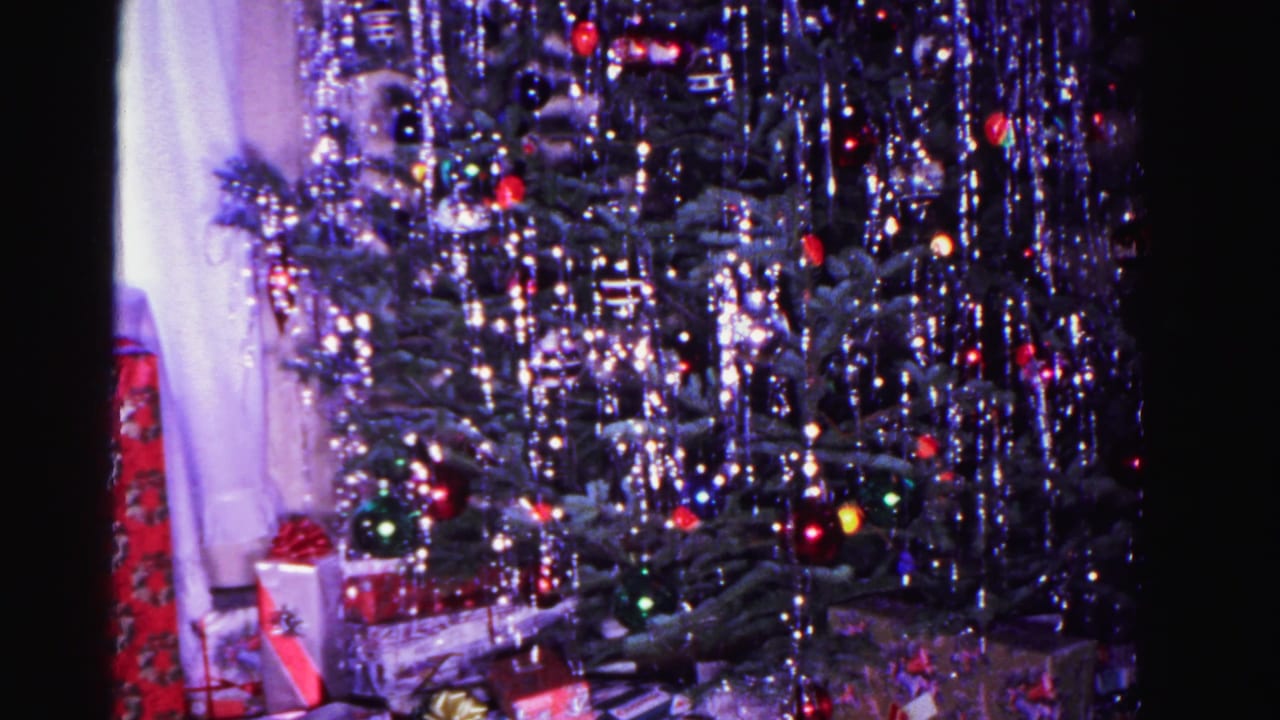 8mm home movie frame circa 1962: tinsel-draped Christmas tree with multicolored lights and holiday gifts in a domestic interior, sheer curtain at right edge.