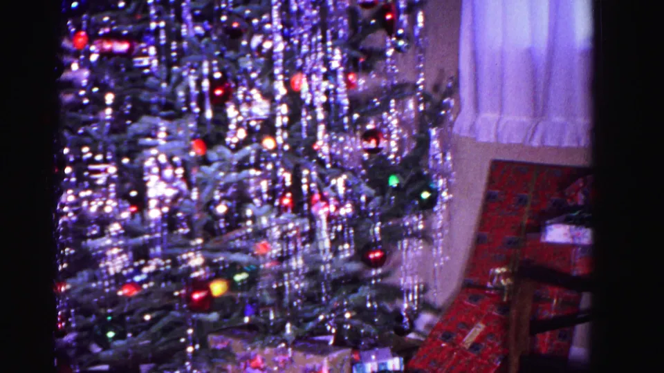 8mm home movie frame circa 1962: Christmas tree with icicle tinsel and colored ornament lights; upholstered chair and sheer curtain visible at right edge of domestic interior.