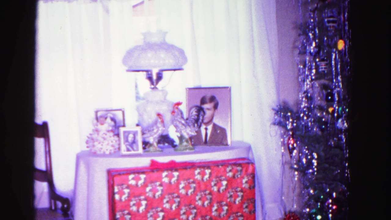 White-draped display table with glass lamp, framed portrait, ceramic roosters, and red poinsettia gift wrap, beside a tinsel-covered Christmas tree.
