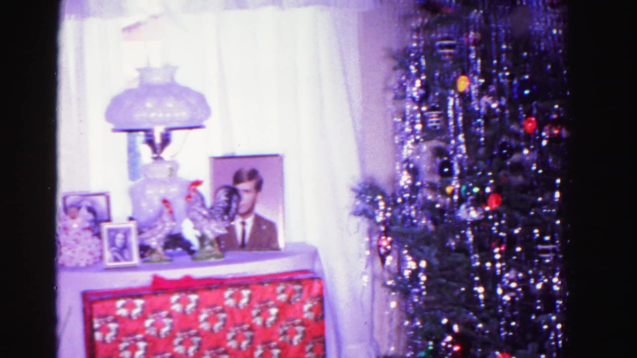 Early-1960s American living room; display table holding a glass lamp, framed adult male portrait, and ceramic roosters adjacent to a Christmas tree heavy with silver tinsel and round ornaments.