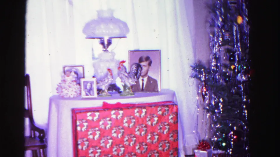 Interior of a 1962 American home at Christmas: table with red holiday cloth, framed portrait photograph of a man, glass globe lamp, and a tinsel tree with colored lights at right.