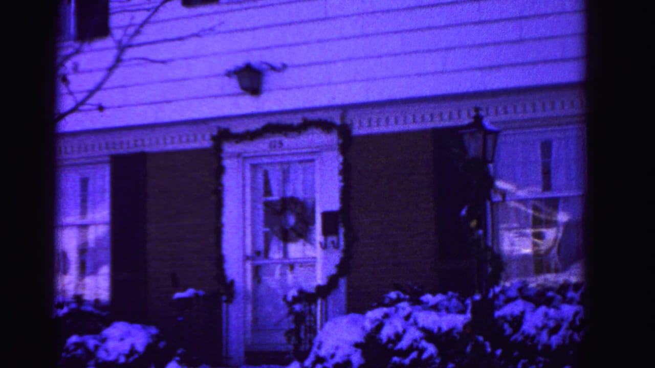 Two-story suburban house with brick lower facade and white clapboard siding, Christmas wreath on left window, snow-covered shrubs in foreground, strong purple color cast.