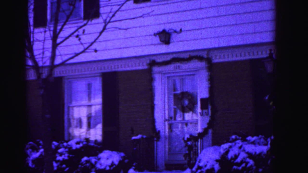 Suburban house front panned slightly right, white siding and brick base, Christmas wreath on right window, dark shutters, snow-covered shrubs, bare tree at edge, purple film cast.