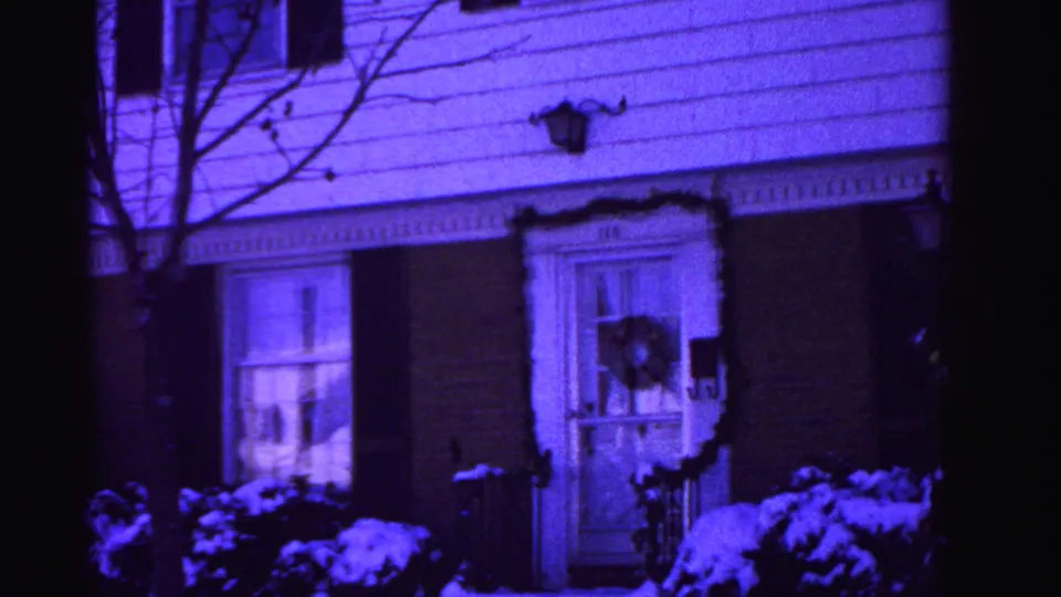 8mm home movie frame circa 1962 showing a Hagerstown house panned slightly right with Christmas wreath, dark shutters, snow-covered shrubs, bare deciduous tree, and pronounced purple film-aging cast.