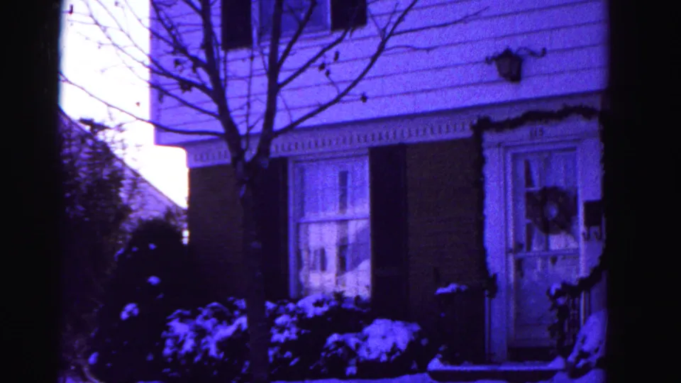 8mm home movie frame circa 1962 centering on the front door of a Hagerstown house, showing a Christmas wreath on the door, lace-curtained windows, brick facade, bare tree, and snow-covered ground.