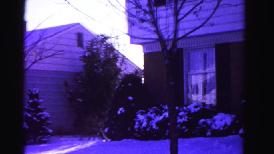 8mm home movie frame circa 1962 at the end of the pan across a Hagerstown suburban house, revealing an adjacent lower structure, snow-covered evergreen shrubs, bare trees, and snow-covered ground.