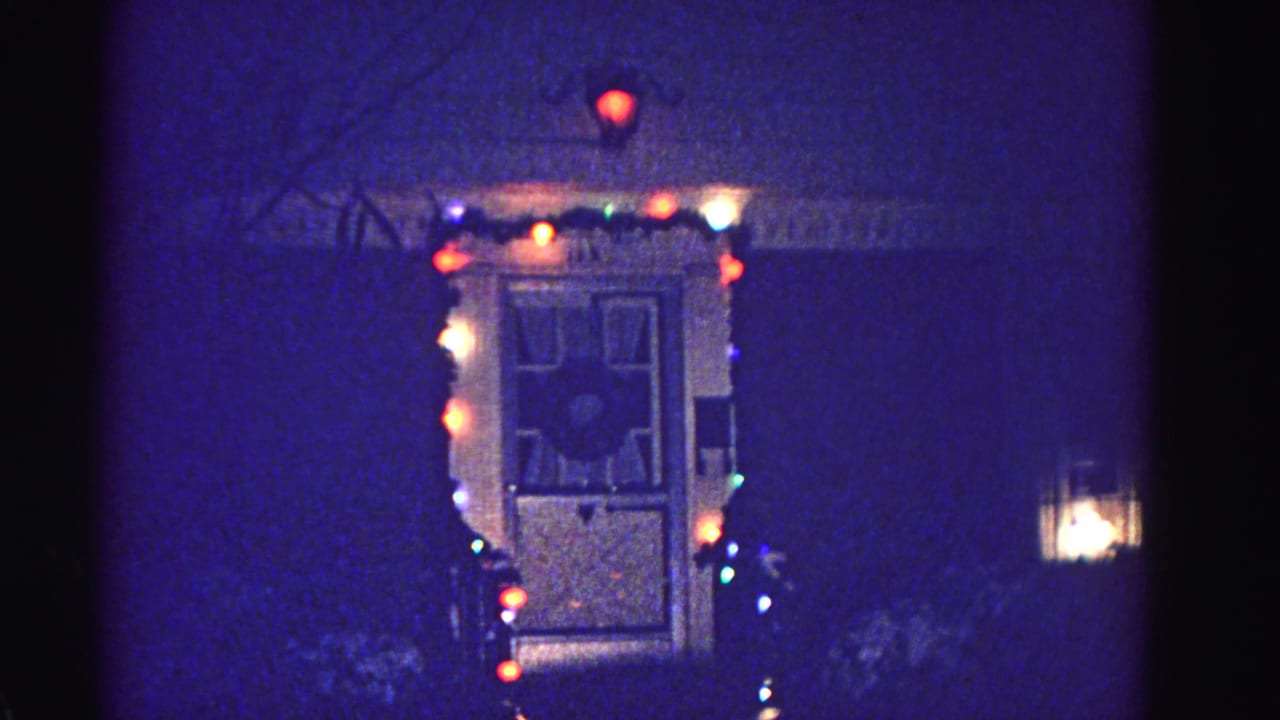 Front door frame strung with multicolored Christmas lights at night; strong blue 8mm film cast and visible grain.