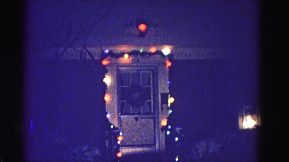 Wood-panel residential door outlined with multicolored Christmas string lights and a warm porch lantern, 8mm night footage, Hagerstown Maryland, circa 1962.