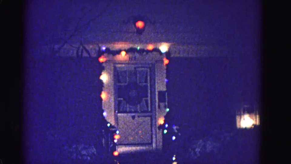 Close view of a panel door outlined in multicolored Christmas bulbs with a warm porch lantern at right, 8mm home movie, Hagerstown Maryland, circa 1962.