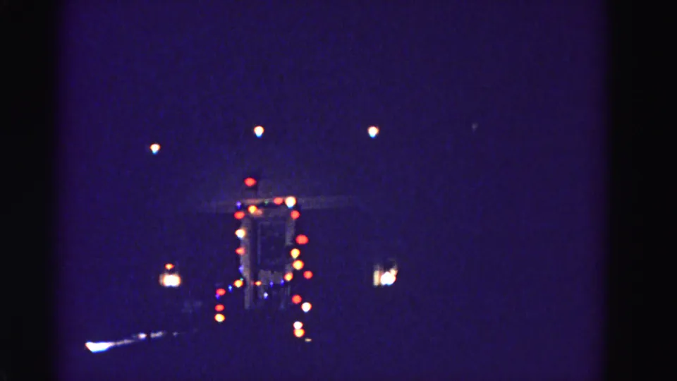8mm home-movie frame circa 1962 showing a distant festively illuminated building at night; camera has shifted slightly, revealing a second lit element at left edge.