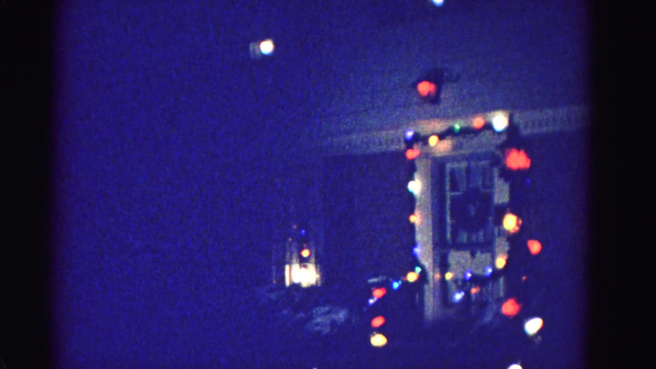 A house front door framed by colorful string lights with a hanging lantern to the left, filmed at night on 8mm.