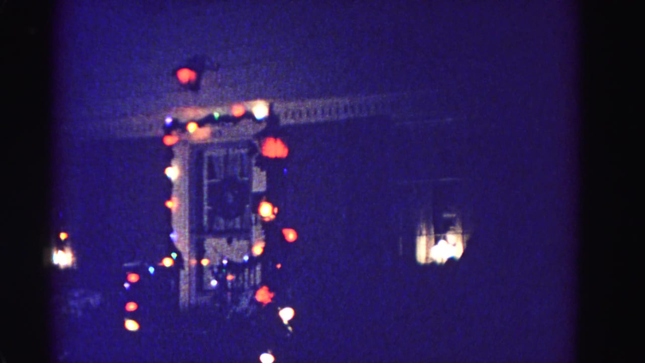 1962 8mm footage of a residential house in Hagerstown, Maryland decorated with multicolored Christmas lights along roofline and porch, warm interior light visible in window.