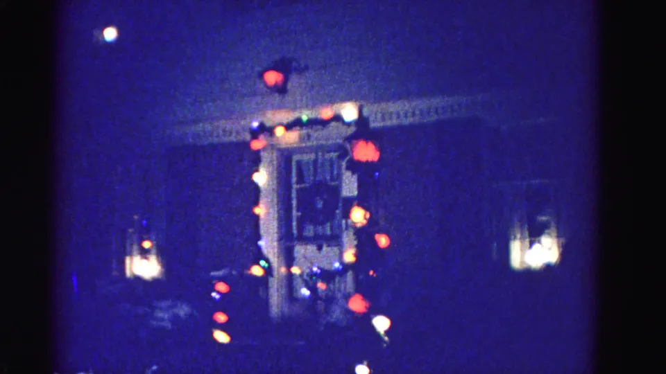 Mid-clip frame from a 1962 8mm home movie showing a Christmas-lit house exterior in Hagerstown, Maryland with an exterior lantern fixture and lit window.