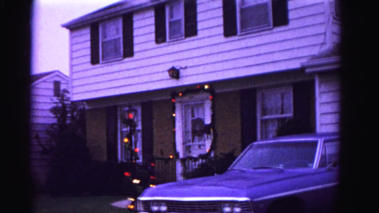 Two-story white-sided house with black shutters, Christmas string lights on porch, and an early-1960s American sedan in the driveway; blue-purple 8mm color cast.