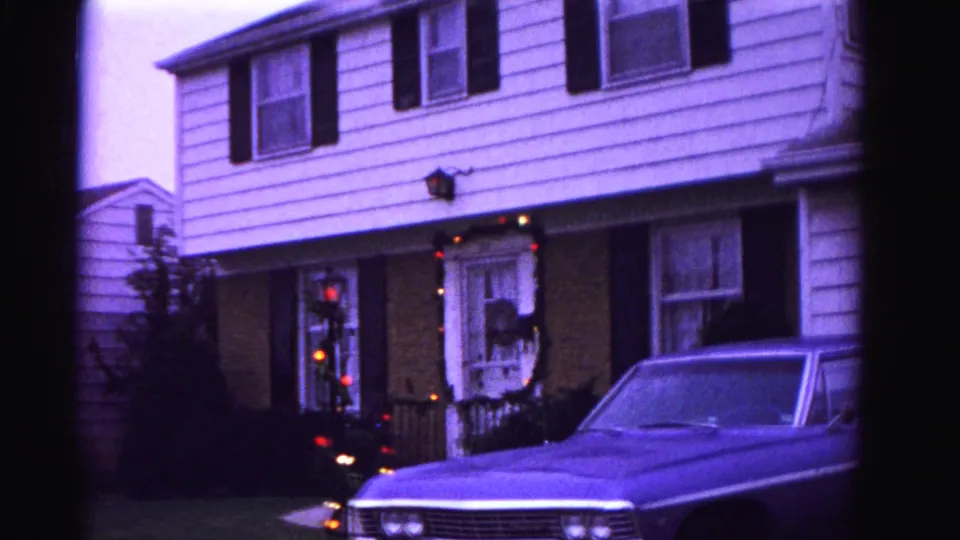 Suburban two-story house in Hagerstown, Maryland, circa 1962, with multicolored Christmas string lights on porch and door; large American sedan in driveway; pronounced blue-purple 8mm color cast.