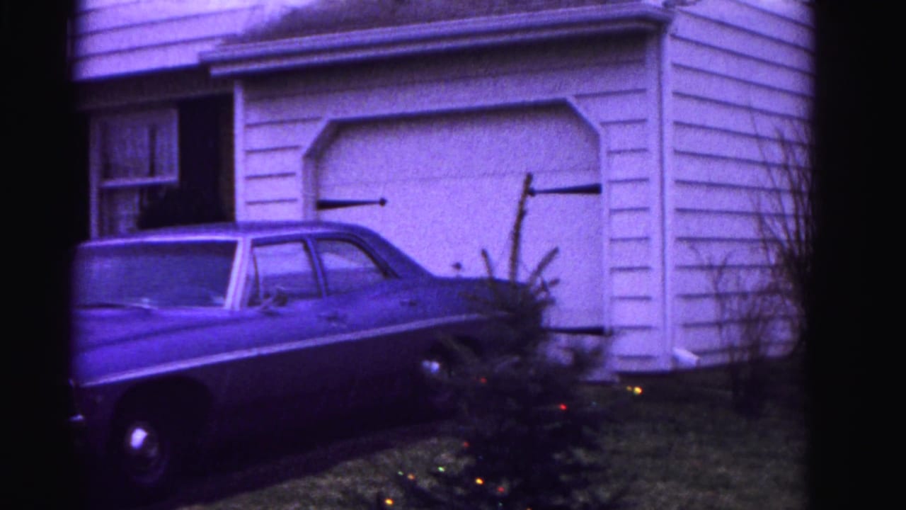 8mm home movie frame, ca. 1962, Hagerstown Maryland: full-size American sedan beside a white clapboard house; Christmas-decorated small evergreen tree in yard; strong purple film cast.