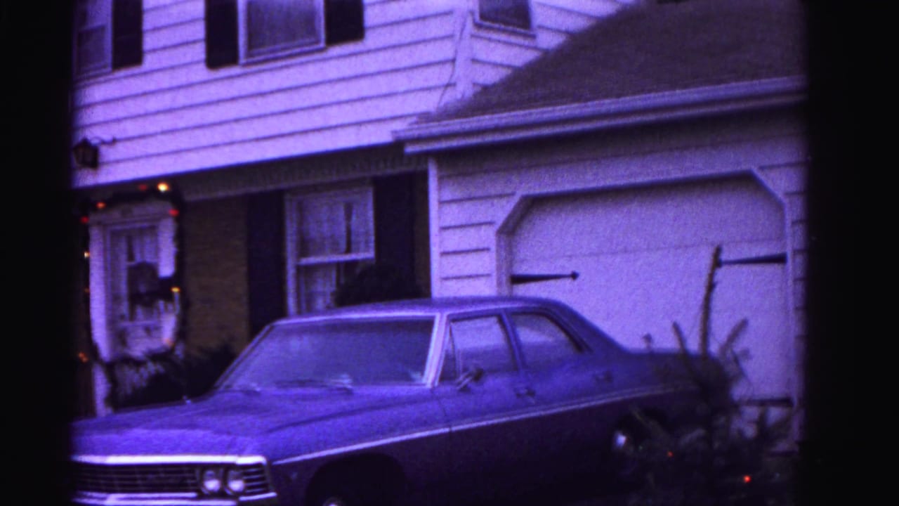 8mm home movie frame, ca. 1962, Hagerstown Maryland: two-story house facade with black shutters, Christmas wreath and garland at front entrance, full-size sedan in driveway; purple-shifted color.