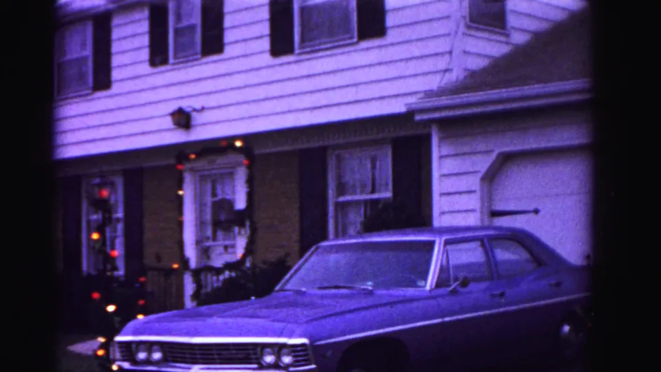 8mm home movie frame, ca. 1962, Hagerstown Maryland: car front end with dual headlights in foreground; Christmas wreath and glowing string lights on doorway; exterior wall sconce; aged purple-blue cast.