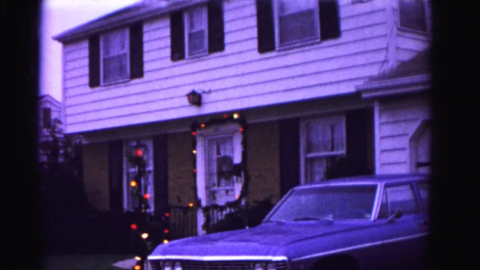 8mm home movie frame, ca. 1962, Hagerstown Maryland: car hood in foreground, Christmas wreath and garland on lit doorway, wall sconce illuminated; purple-blue 8mm color cast throughout.