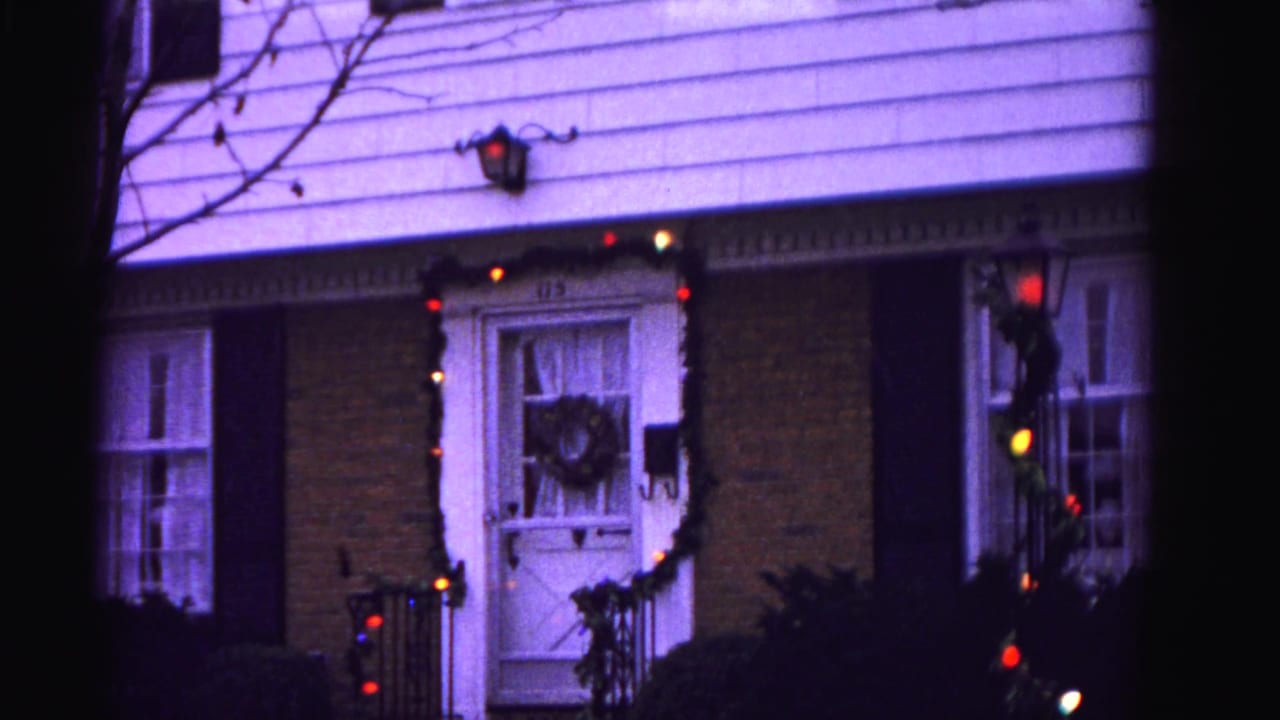 1962 suburban home in Hagerstown, Maryland — brick facade with black shutters, Christmas wreath on front door, and multicolored garland lights, 8mm purple color cast.