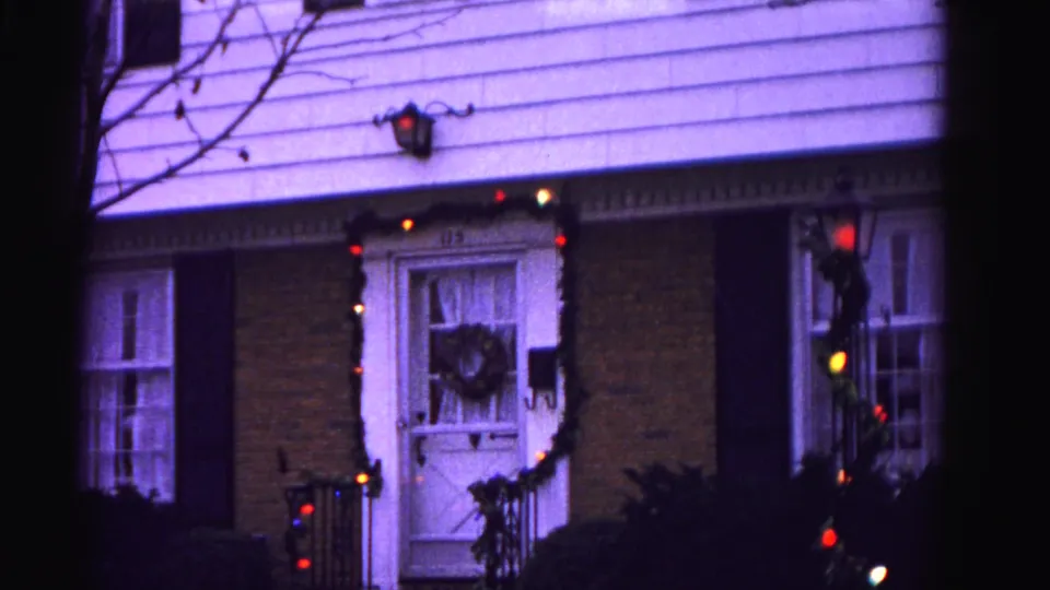 1962 suburban home in Hagerstown, Maryland — brick facade with black shutters, Christmas wreath on front door, and multicolored garland lights, 8mm purple color cast.