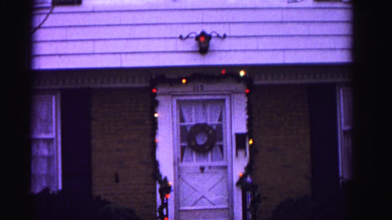 White front door with wreath and house number 115, framed by colored Christmas lights and garland, wall lantern above, brick and clapboard exterior at dusk.