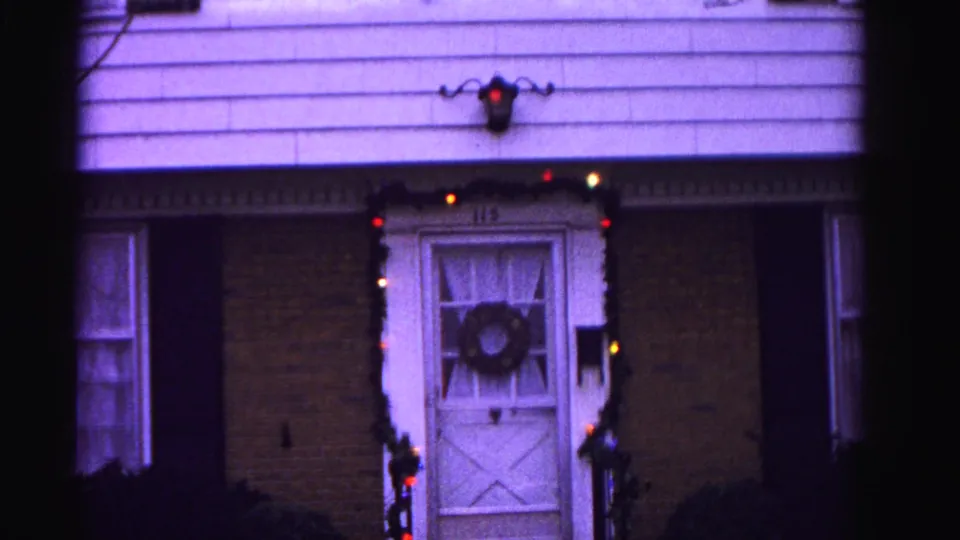 8mm film frame circa 1962 showing the decorated front entrance at house 115, Hagerstown, Maryland — wreath on door, multicolored lights, and glowing wall lantern.