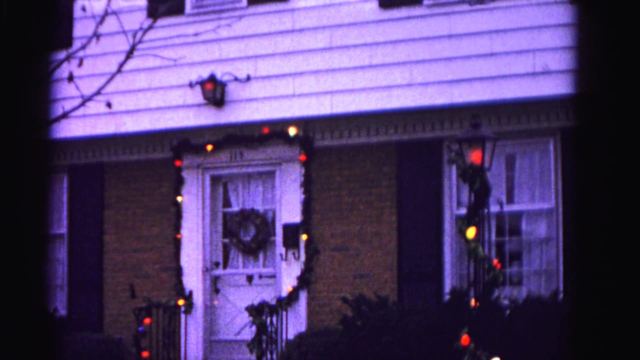 8mm frame circa 1962, Hagerstown, Maryland — front door with wreath, house number 115, red and yellow Christmas lights strung across the entrance, wall lantern glowing.