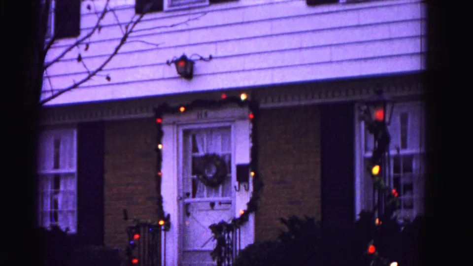 Final 8mm frame circa 1962, Hagerstown, Maryland — Christmas-decorated front entrance with colored light bulbs, glowing wall lantern, wreath on door, and porch-level shrubbery.