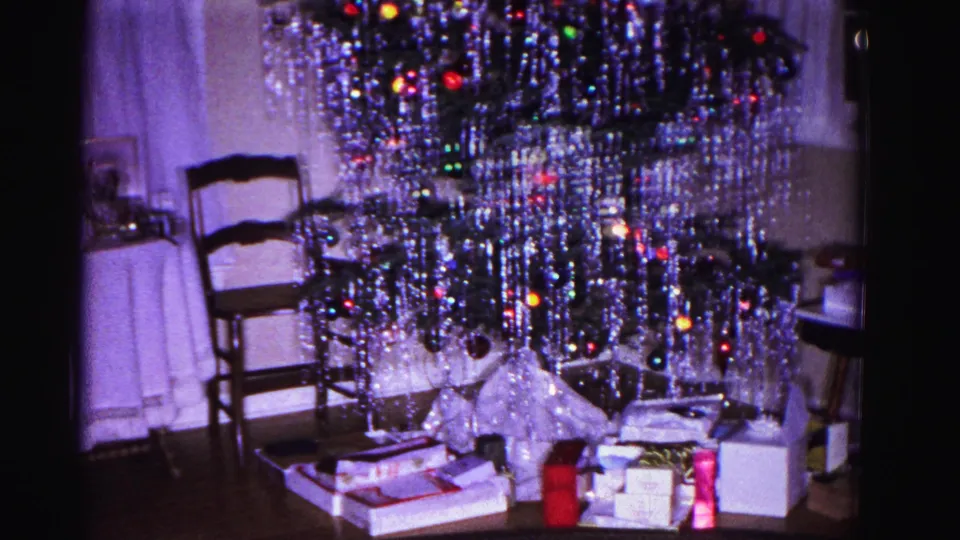 1962 8mm home-movie interior: tinsel-covered Christmas tree with colorful incandescent lights and gift boxes beneath, wooden rocking chair at left, Hagerstown, Maryland.