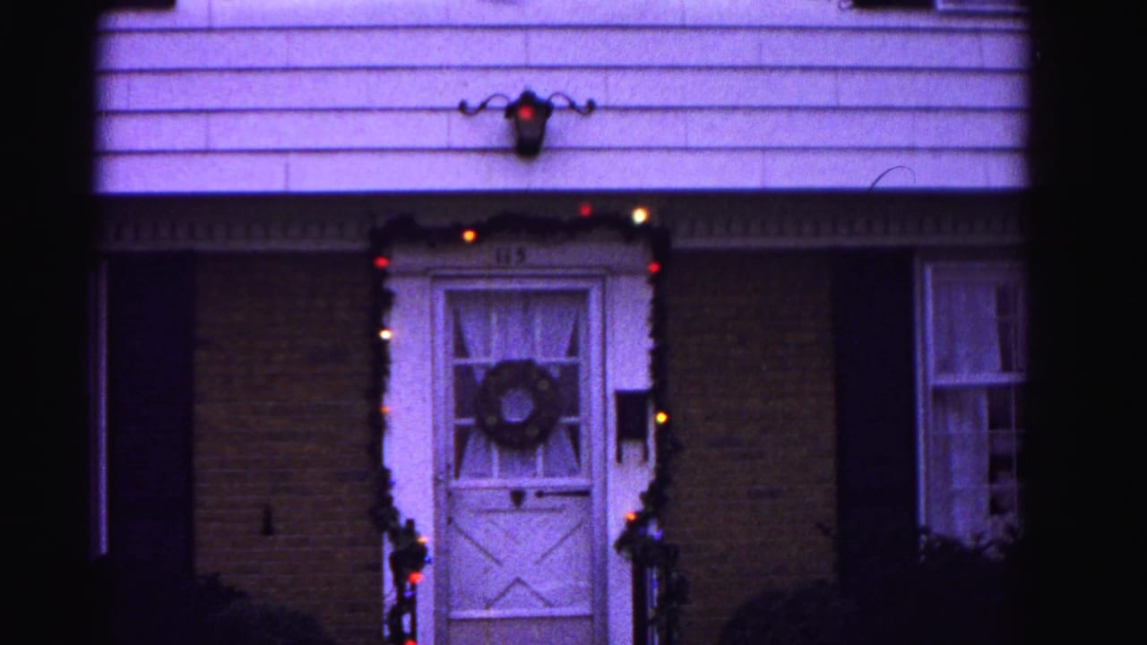 Exterior of brick house with white front door decorated with a wreath and colored string lights framing the doorway; white clapboard upper siding and wall-mounted lantern.